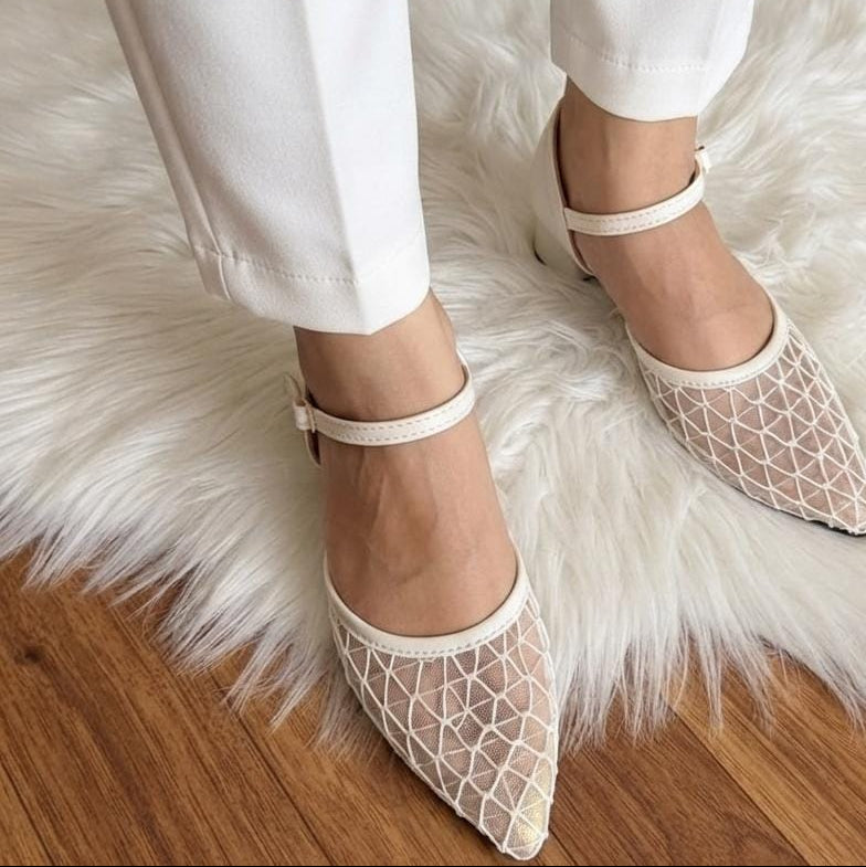 A person wearing white pointed-toe flats with a geometric openwork mesh pattern and ankle straps, standing on a white faux-fur rug over a wood floor.