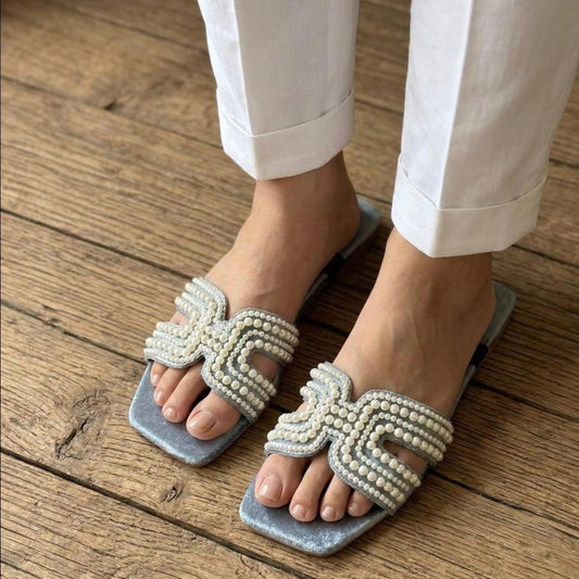 A close-up of a person wearing white pants and silver flat slide sandals with ornate pearl-embellished straps, standing on a rustic wooden plank floor.