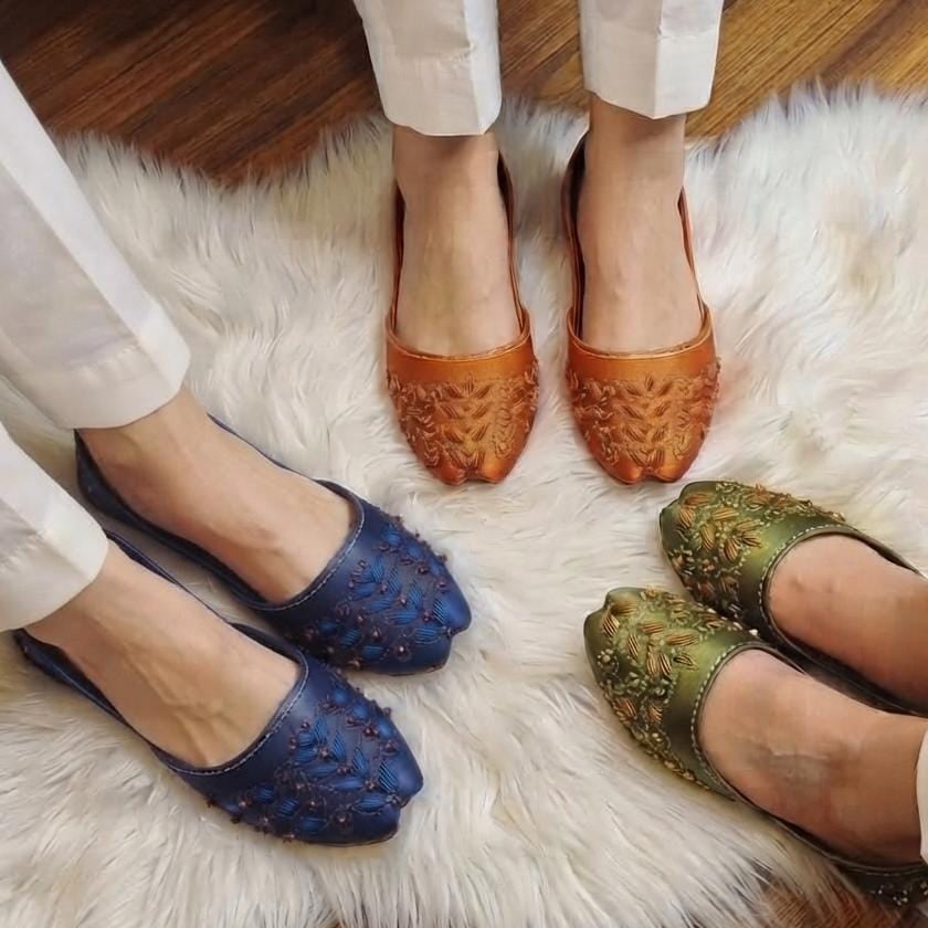 Three pairs of women's flat shoes with textured, studded leather uppers are shown on feet wearing white trousers, arranged on a white faux fur rug over a wooden floor. The shoes are in blue, brown, and olive green.