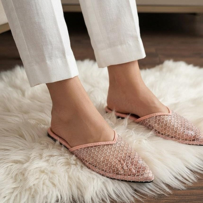 A person wearing light pink pointed-toe mule flats with a textured geometric mesh pattern, styled with white trousers on a white faux fur rug over a wooden floor.