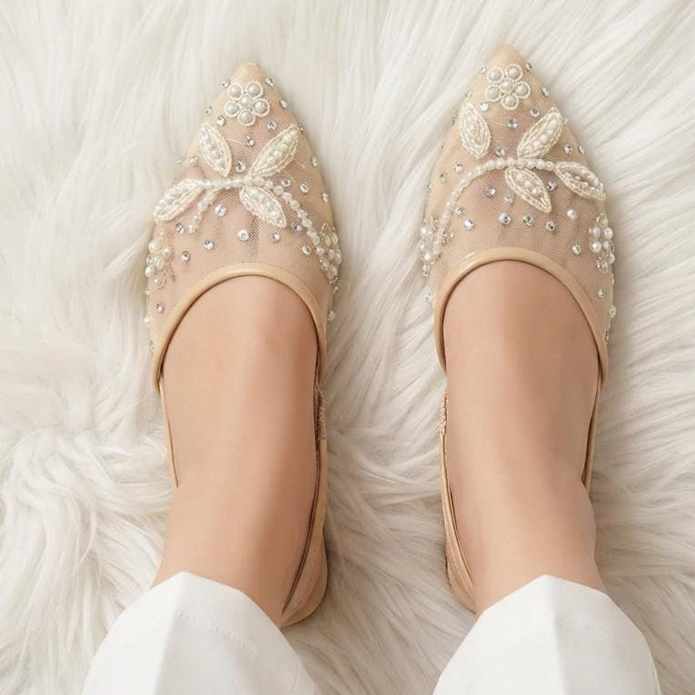 A pair of beige pointed-toe flats decorated with intricate white floral beadwork and rhinestone embellishments, shown on a model's feet resting on a white faux fur rug.