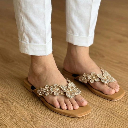 Brown sandals with floral embellishments worn with white pants on a wooden floor.
