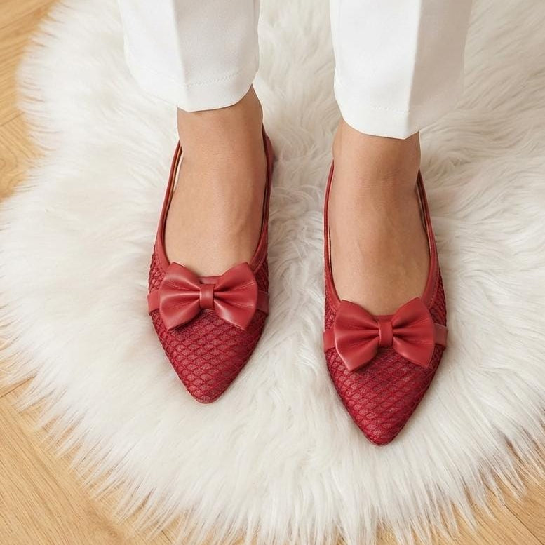 A pair of red pointed-toe flats with a textured woven pattern and a matching fabric bow on the toe, shown on a model wearing white pants while standing on a white faux fur rug.