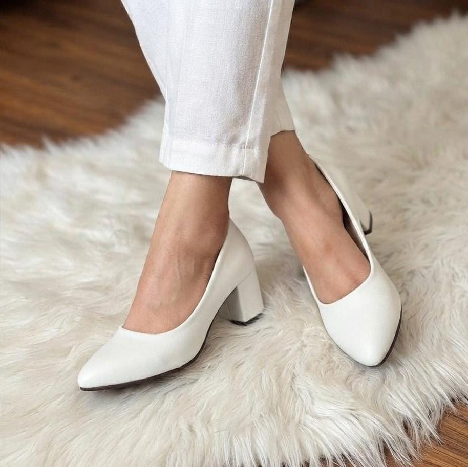 A person wearing white pointed-toe block-heel pumps with white cropped pants, standing on a white faux-fur rug over a wooden floor.