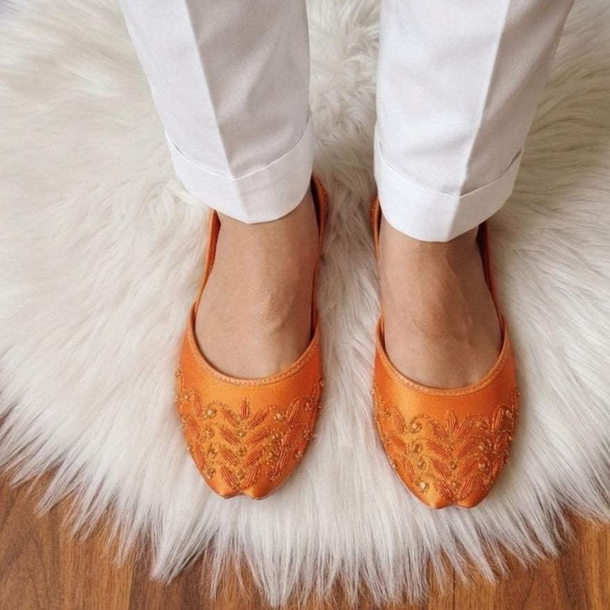 A person wearing white pants and bright orange pointed flats with gold embroidery, standing on a white faux-fur rug over a wooden floor.