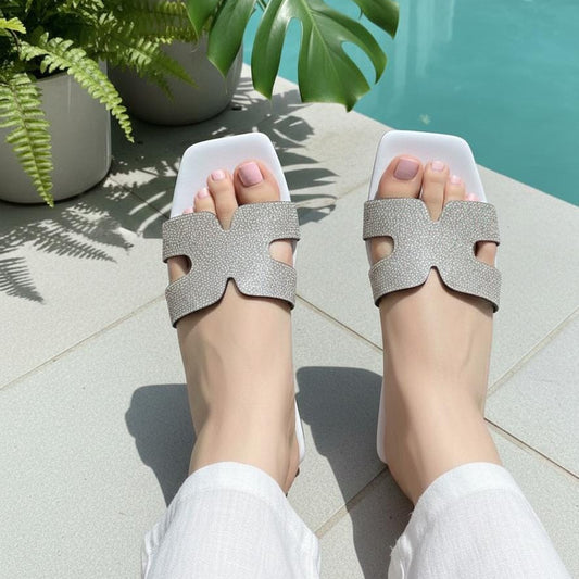A person wearing white slide sandals with wide glittery silver straps, photographed from above near a pool with green plants in the background.