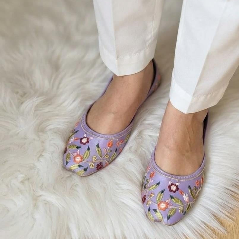 A person wearing white pants and lavender slip-on flats with colorful floral embroidery, standing on a white faux-fur rug.