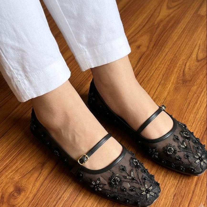 Black embellished shoes worn with white pants on a wooden floor