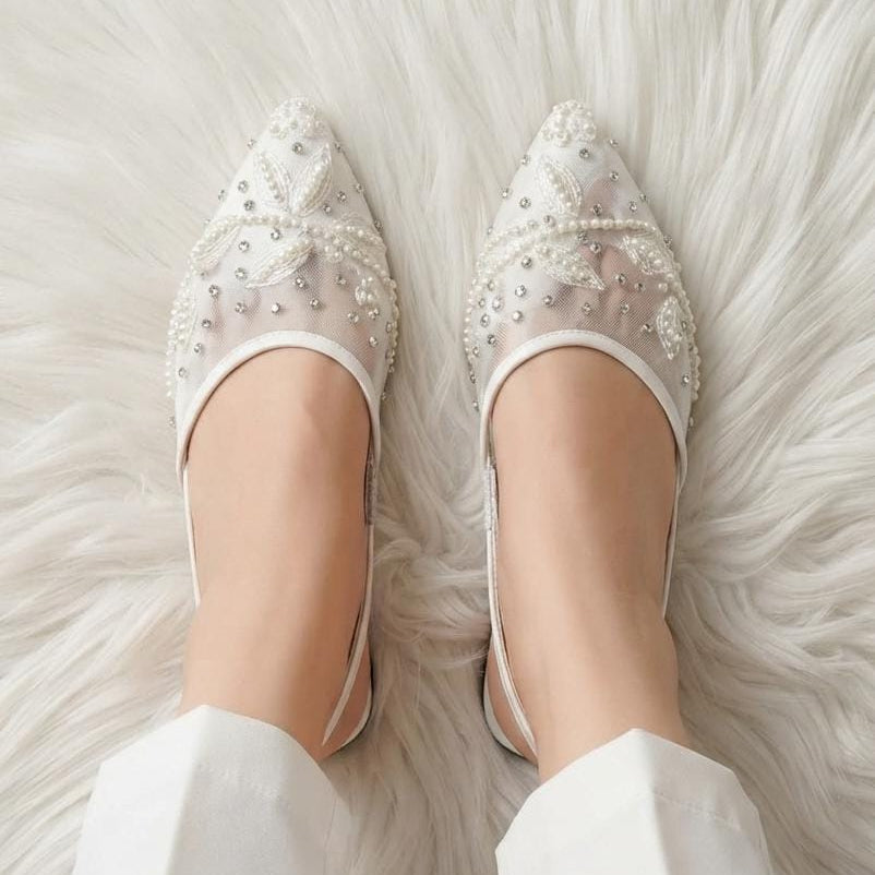 A pair of white pointed-toe slingback flats decorated with pearls and rhinestones, shown on a person's feet resting on a white faux fur rug.