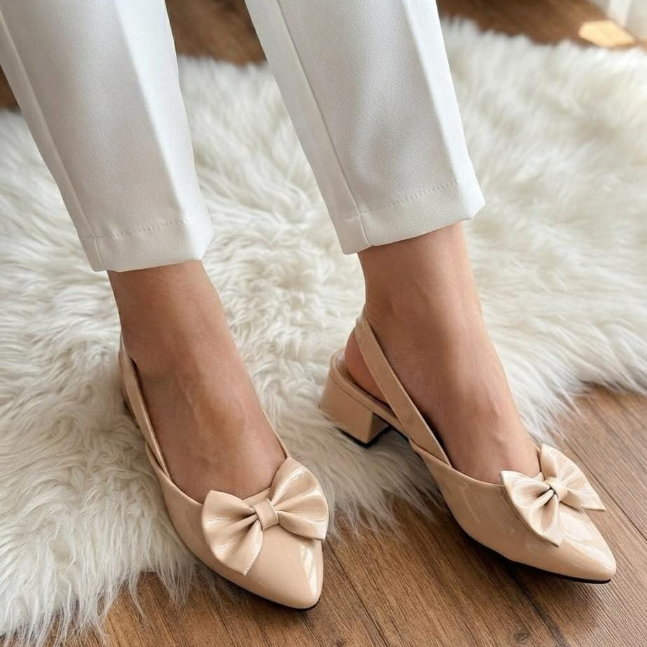 A person wearing beige slingback low-heel pumps with large bow details on the toes, paired with white pants, standing on a white faux fur rug over a wooden floor.