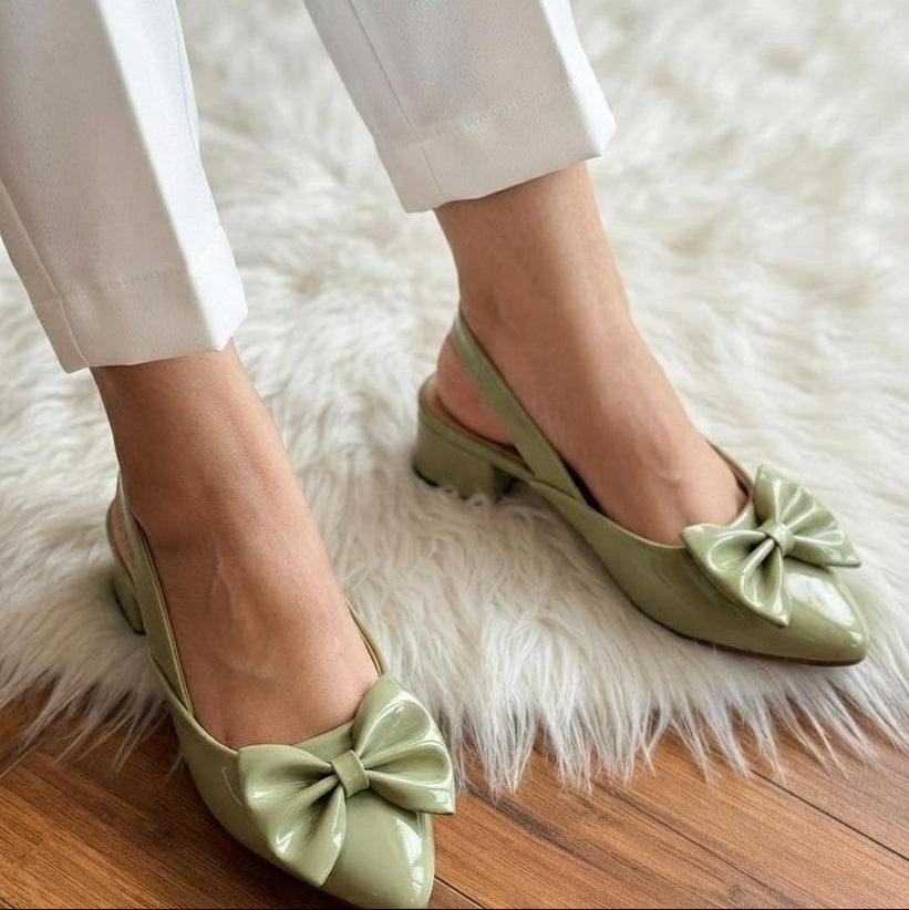 A person wearing light sage green slingback pointed-toe flats with large bow details, standing on a white faux-fur rug over a wooden floor.