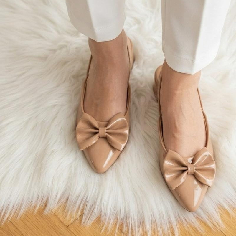 A person wearing beige patent leather ballet flats with large bow details, standing on a white faux fur rug over a light wood floor.