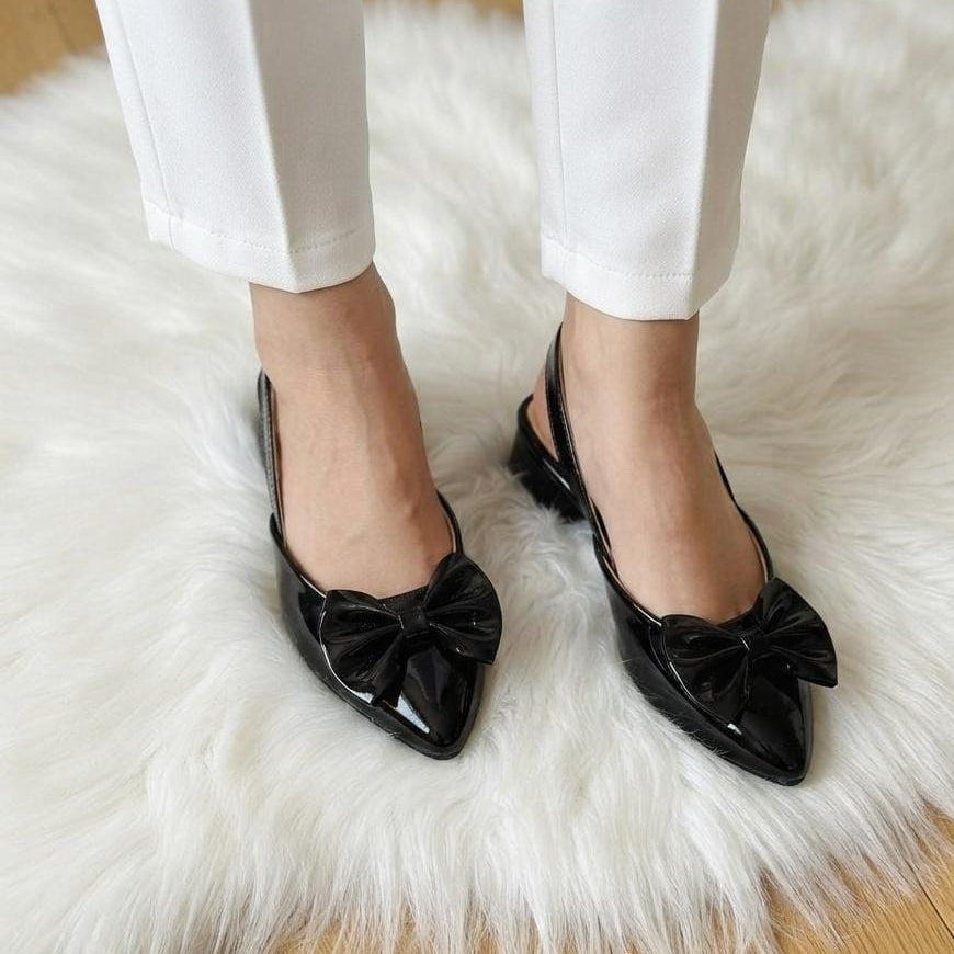 A person wearing white pants and black patent leather slingback flats with large bow details on the toes, standing on a white faux fur rug over a light wood floor.