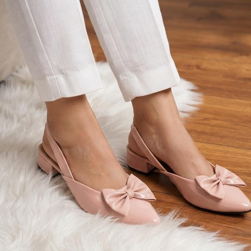A person wearing light pink slingback low-heel flats with bow details, paired with white cropped pants, standing on a white faux fur rug over a wooden floor.