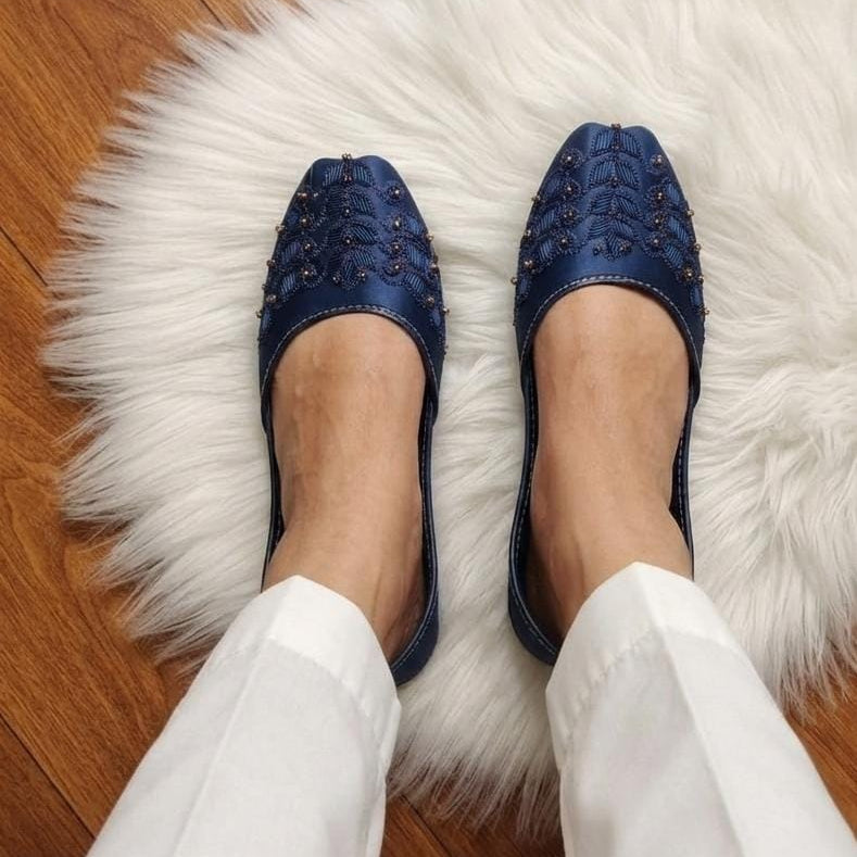 A person wearing blue pointed-toe flats with decorative metallic studs, standing on a white faux-fur rug over a wooden floor.