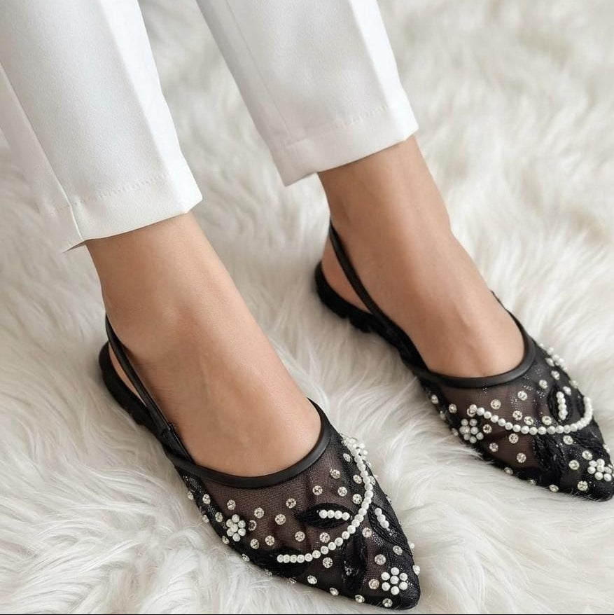 Black pointed slingback flats with sheer mesh and white pearl embellishments, shown on a model wearing white pants on a white faux fur rug.
