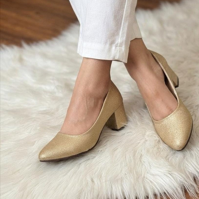 A person wearing pointed-toe heeled pumps in a metallic gold finish, paired with white pants, standing on a white faux fur rug over a wooden floor.