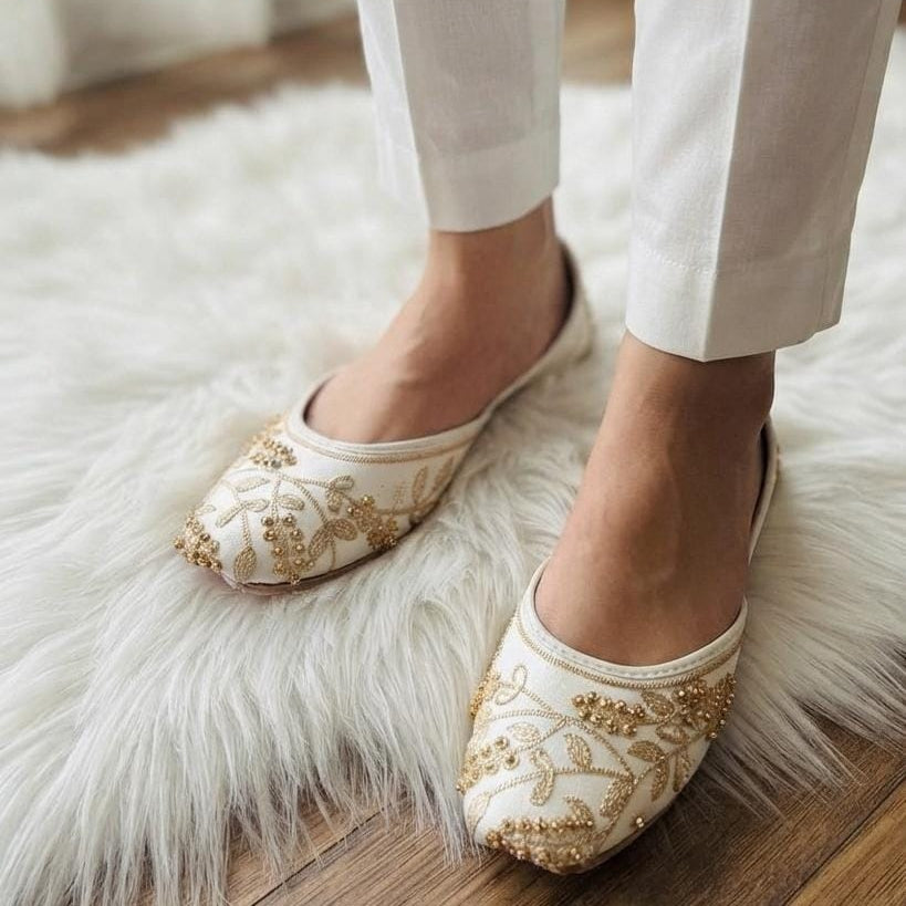A close-up of a person wearing off-white pointed flats with gold floral embroidery and beadwork, standing on a white faux-fur rug over a wooden floor.