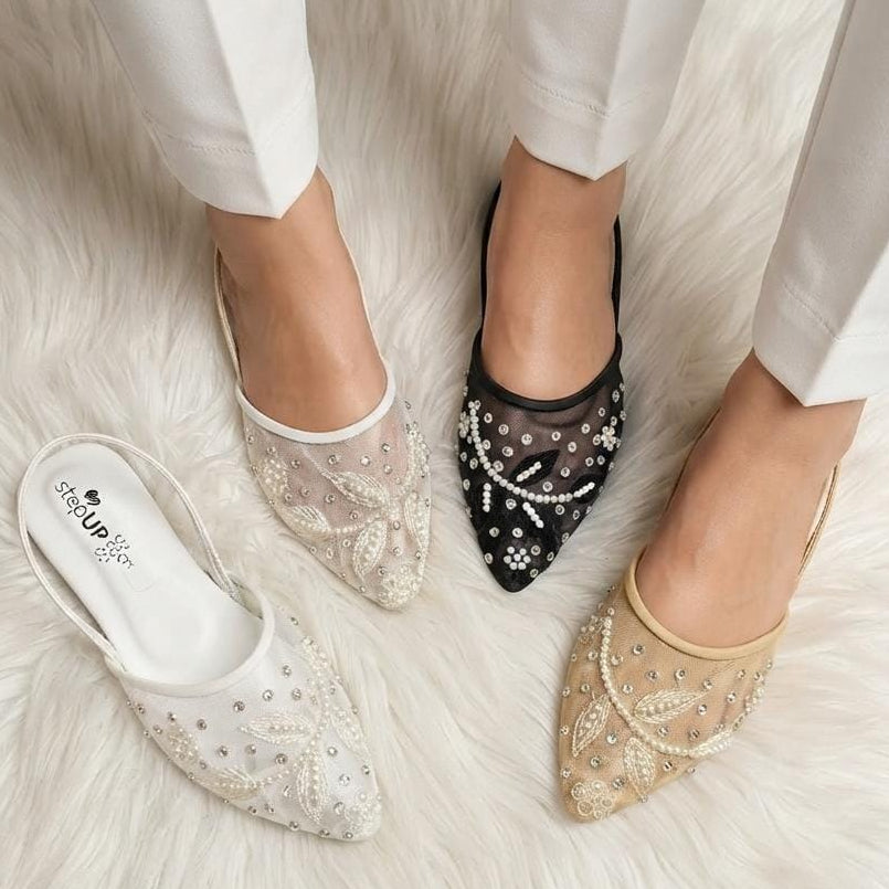 Three embellished slingback pointed-toe flats shown on a white faux-fur surface, with a model's feet wearing two of them. The shoes are in white, black, and beige, each decorated with sheer mesh, pearls, and rhinestone bow details.