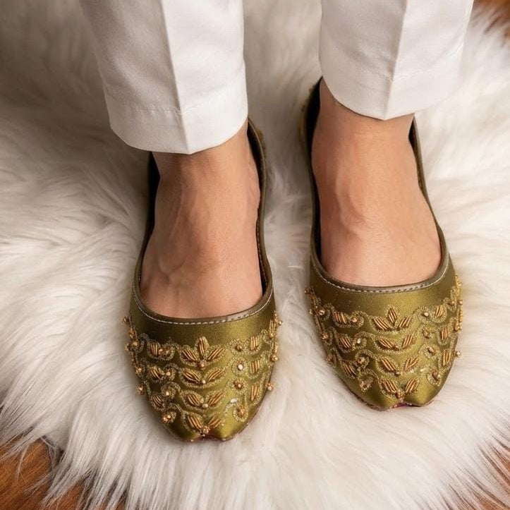 A close-up of a person wearing white pants and olive green pointed-toe flats decorated with gold metallic leaf embroidery, standing on a white faux fur rug.