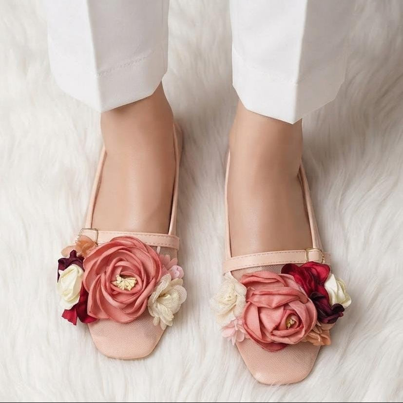 A pair of blush pink flat shoes decorated with large fabric roses in pink, red, cream, and burgundy, shown on a model's feet against a white faux fur background.