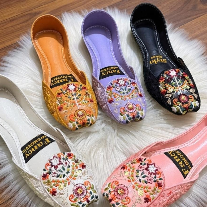 Four slip-on flat shoes with colorful floral embroidery on the toe area, arranged on a white faux fur rug over a wooden floor. The shoes come in white, mustard yellow, lavender, and pink, each with a black insole label reading 'Fancy Shoes'.