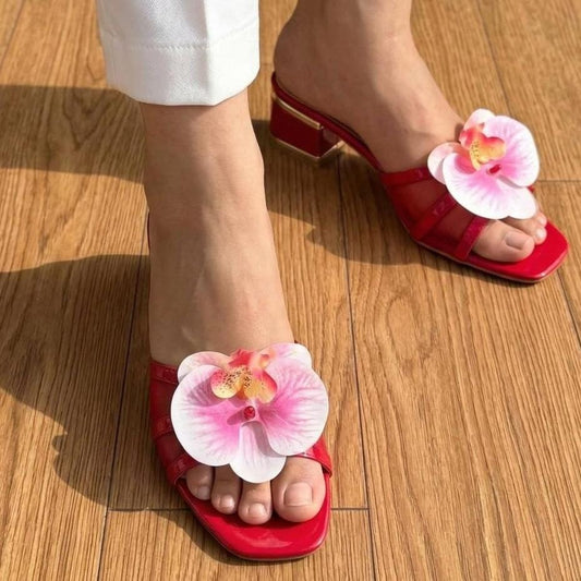 Red open-toe mule sandals with large pink and white orchid flowers on the toes, shown on a model standing on a wooden floor.