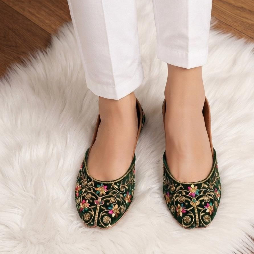 A person wearing dark green embroidered flat shoes with colorful floral patterns, paired with white pants, standing on a white faux fur rug.