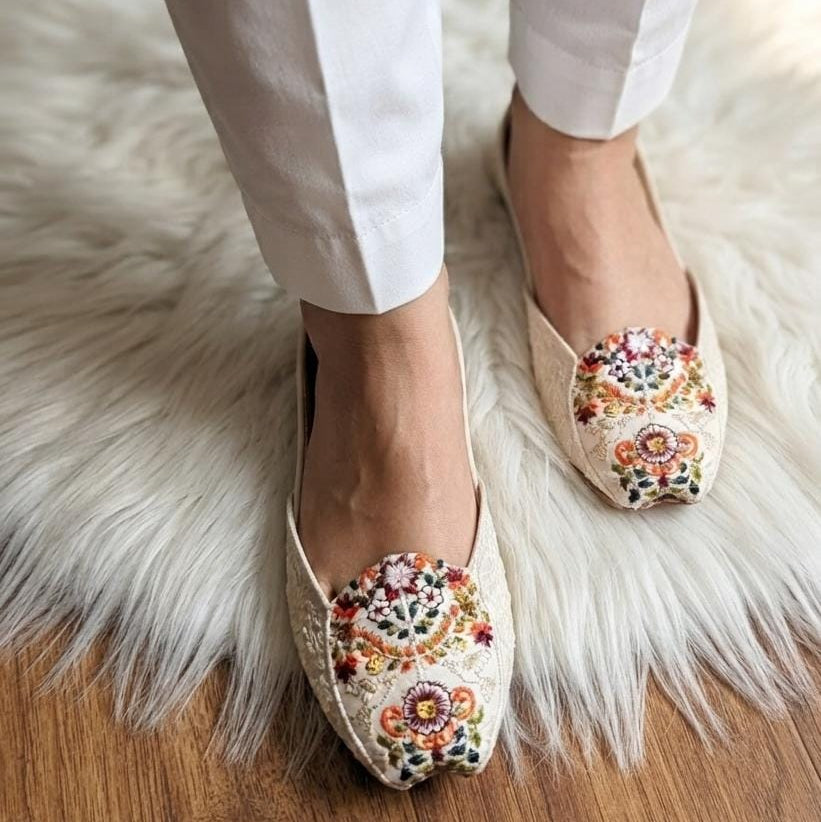 A person wearing white trousers and beige lace flat shoes with colorful floral embroidery on the toes, standing on a white faux-fur rug over a wooden floor.