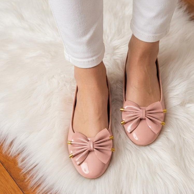 A person wearing white pants and light pink patent leather flats with large bow details and gold-tone studs, standing on a white faux fur rug over a wooden floor.