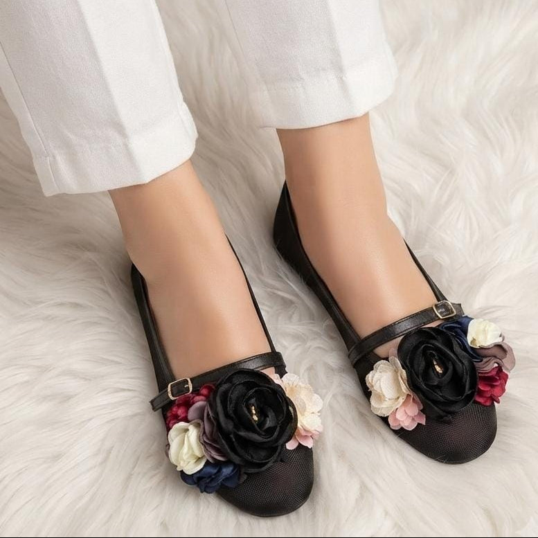Black flat shoes with floral embellishments, worn with white cropped pants on a white faux fur surface.