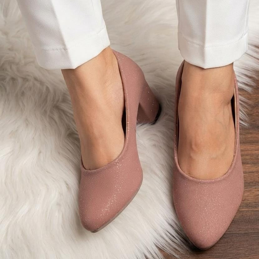 A pair of dusty rose pink glittery block-heel pumps with a caged open-toe design, shown on a model wearing white trousers on a white faux fur rug.
