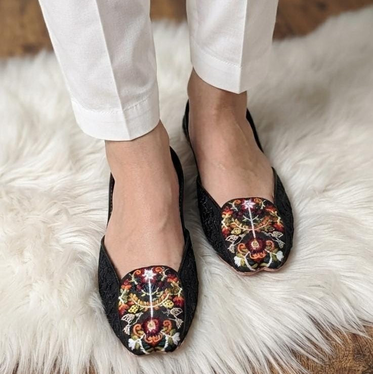 Black lace flat shoes with colorful floral embroidery on the toe, worn with white pants on a white faux fur rug.