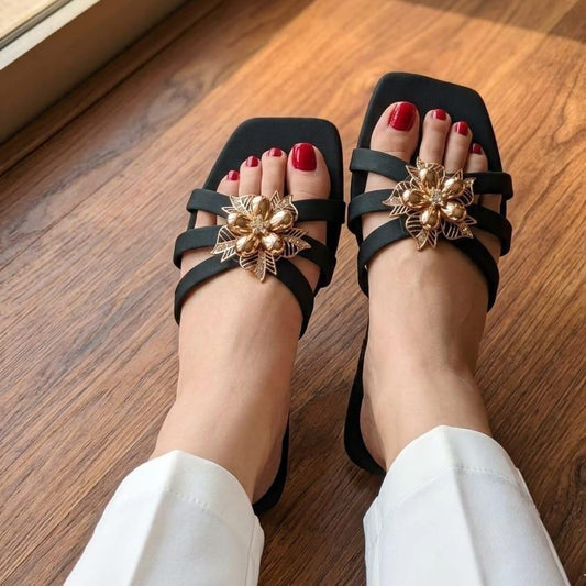 Black strappy flat sandals with a gold-tone floral embellishment on the toes, shown on feet with red toenail polish on a wooden floor.