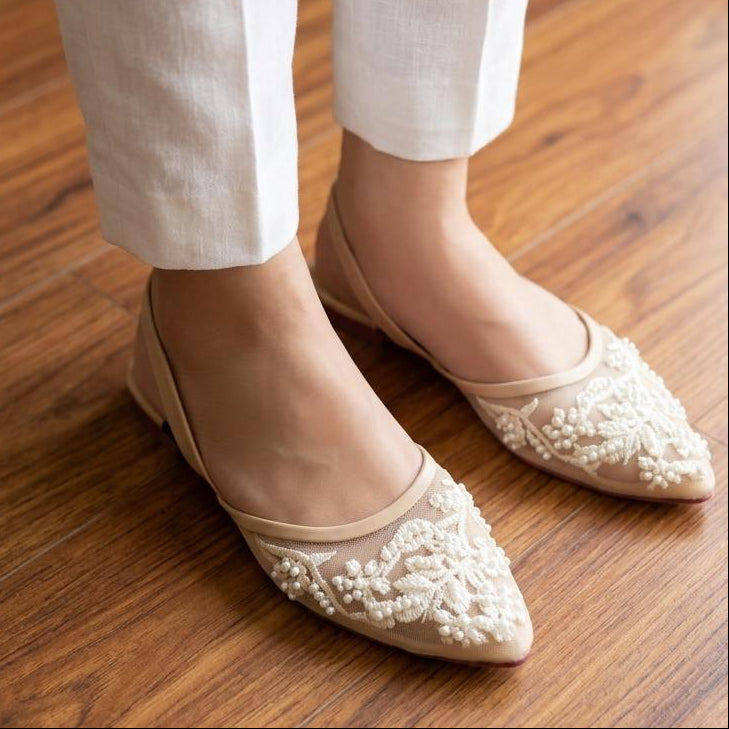 Beige pointed-toe flats with floral embroidery worn with white pants on a wooden floor.