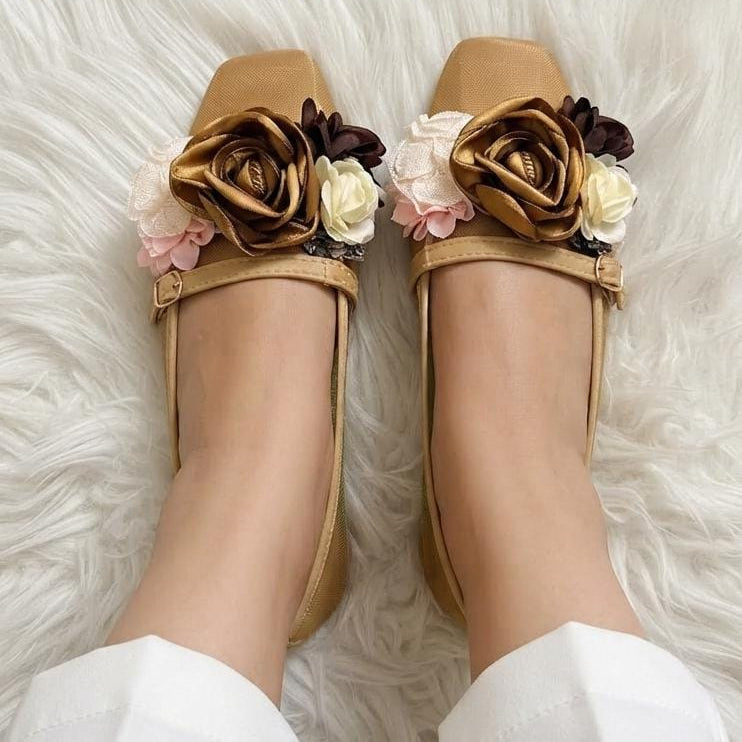 A pair of tan flat shoes with decorative fabric flowers in brown, cream, and pink, worn on feet over a white faux fur rug.