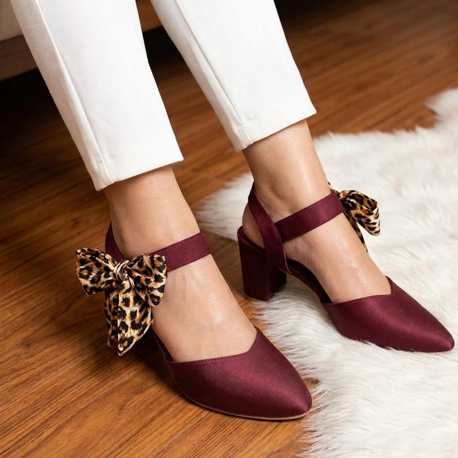 Maroon shoes with leopard print bows worn by a person on a wooden floor.