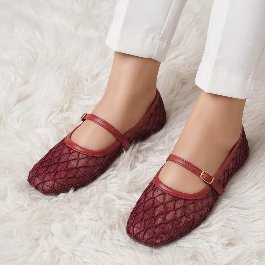 A pair of burgundy crocodile-embossed flat shoes with Mary Jane straps and gold buckle details, shown on a model wearing white pants on a white faux fur rug.