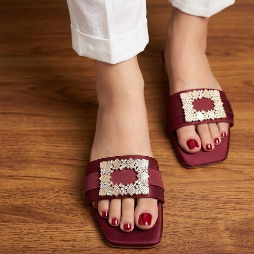 A close-up of a person wearing burgundy slide sandals with ornate white crystal-embellished square straps, paired with white pants, standing on a wooden floor.