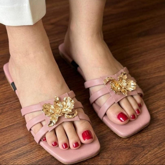 A close-up of a person wearing light pink strappy slide sandals with large gold floral embellishments on the toes, paired with white cropped pants and red toenail polish, standing on a dark wood floor.
