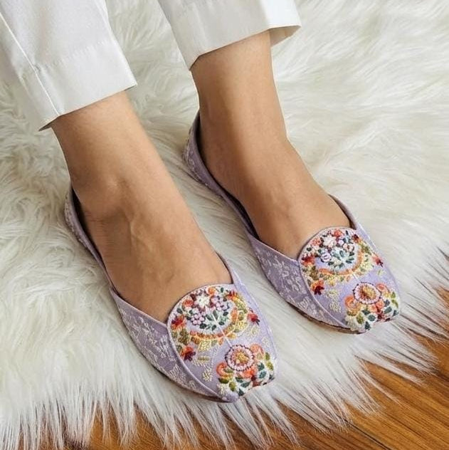 A person wearing light lavender pointed-toe flats with colorful floral embroidery on the toes, standing on a white faux-fur rug over a wooden floor.