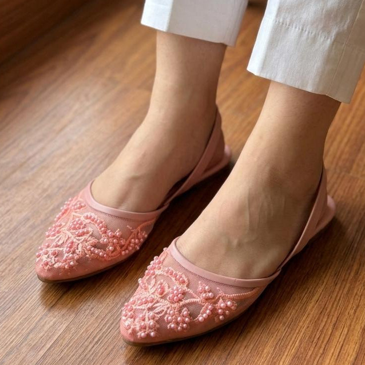 Pink embellished flats worn with white pants on a wooden floor.