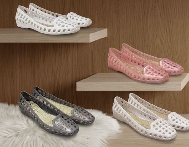 Four pairs of jelly slip-on shoes with perforated cutouts, shown in white, pink, and gray. Two pairs are displayed on light wood floating shelves, and one pair is on a white faux fur rug against a wood-paneled background.